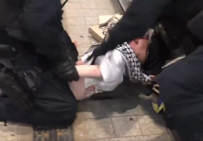 A scene of NSW police brutality raining blows on a young man in a keffiyeh